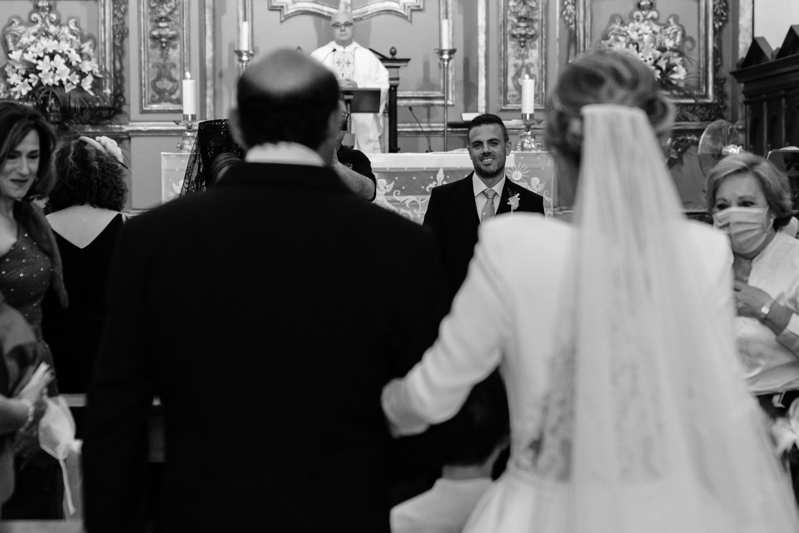 Foto en blanco y negro de la entrada de la novia a la iglesia del brazo de su padre mientras los invitados la miran