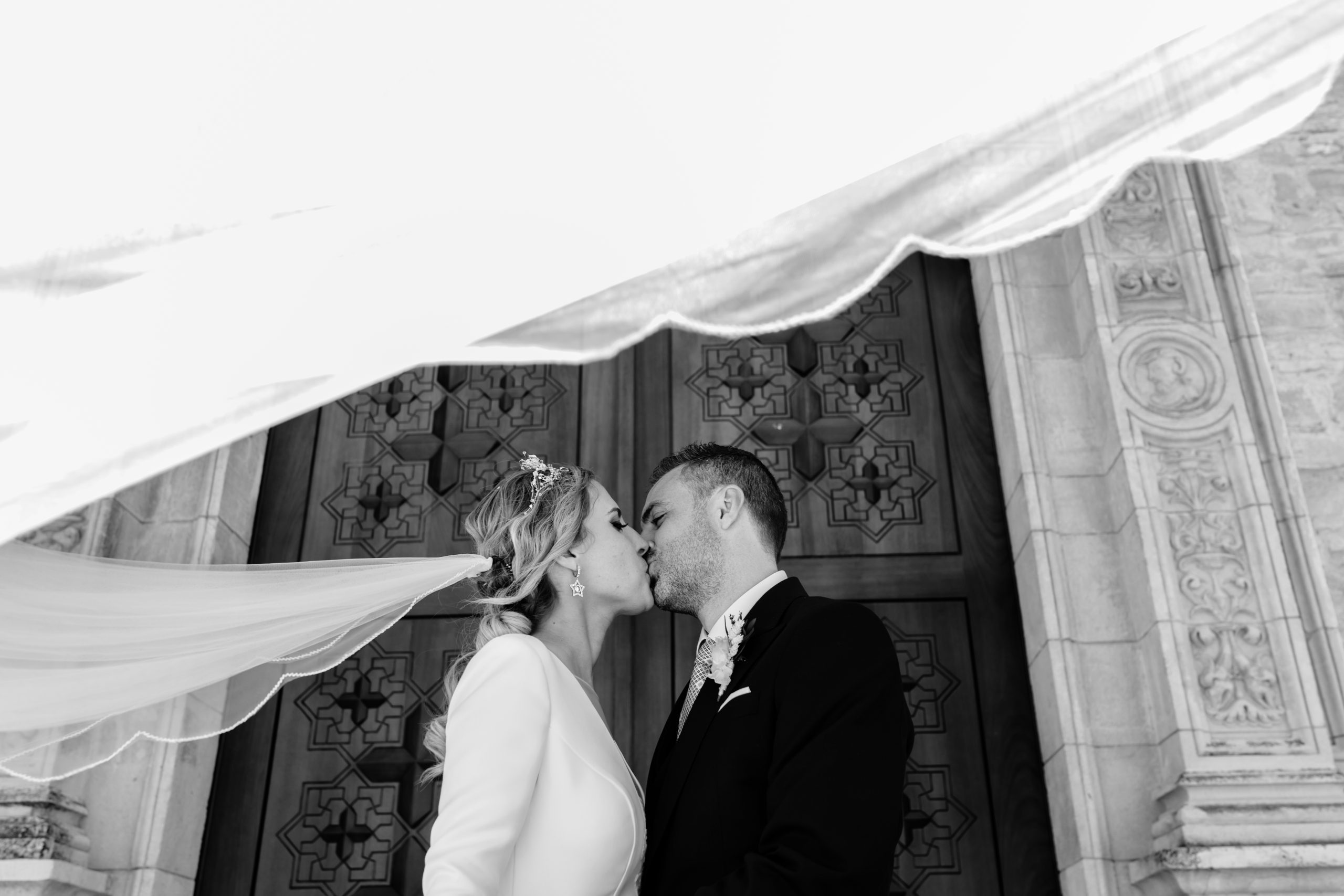 Foto en blanco y negro de los novios besándose delante de una puerta mientras el velo de la novia vuela