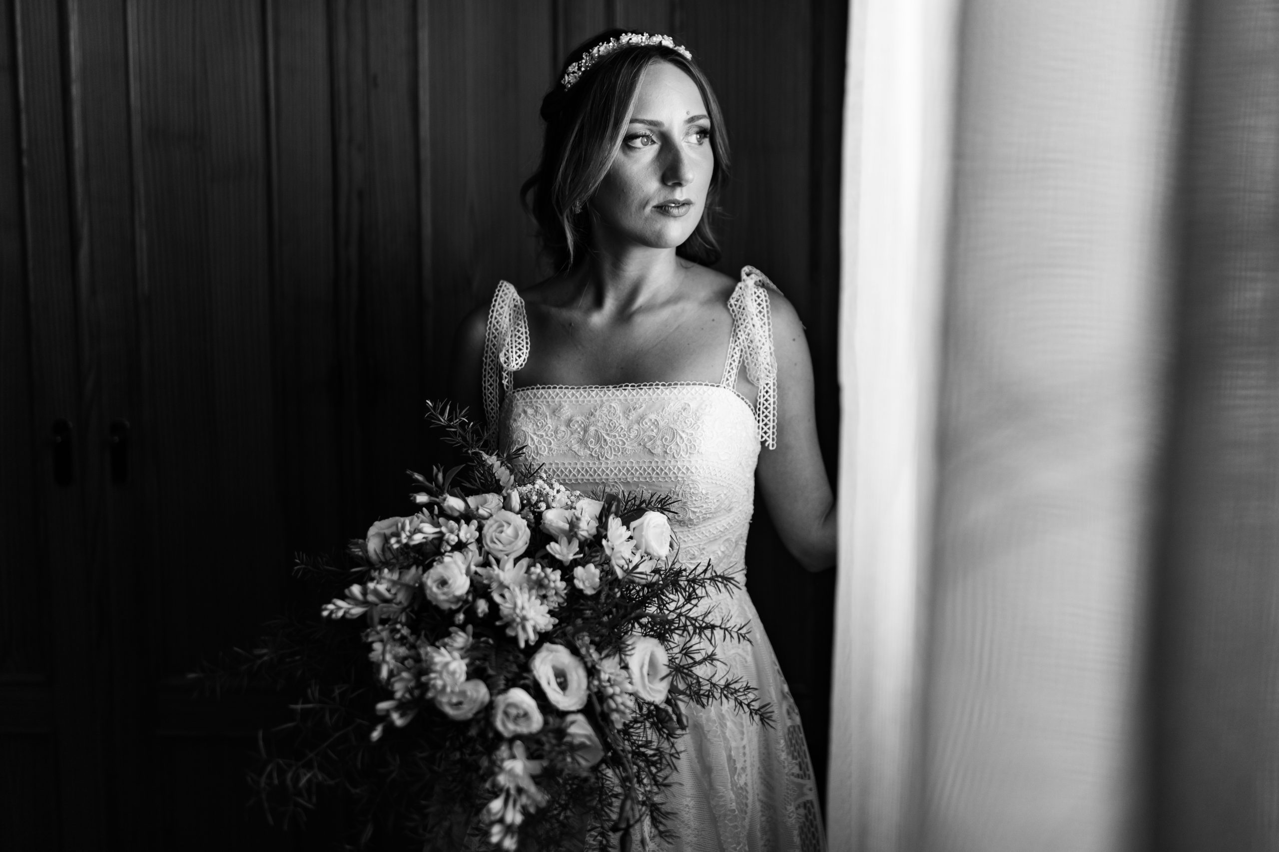 Foto en blanco y negro de la novia preparada con el ramo de flores mirando por la ventana iluminada