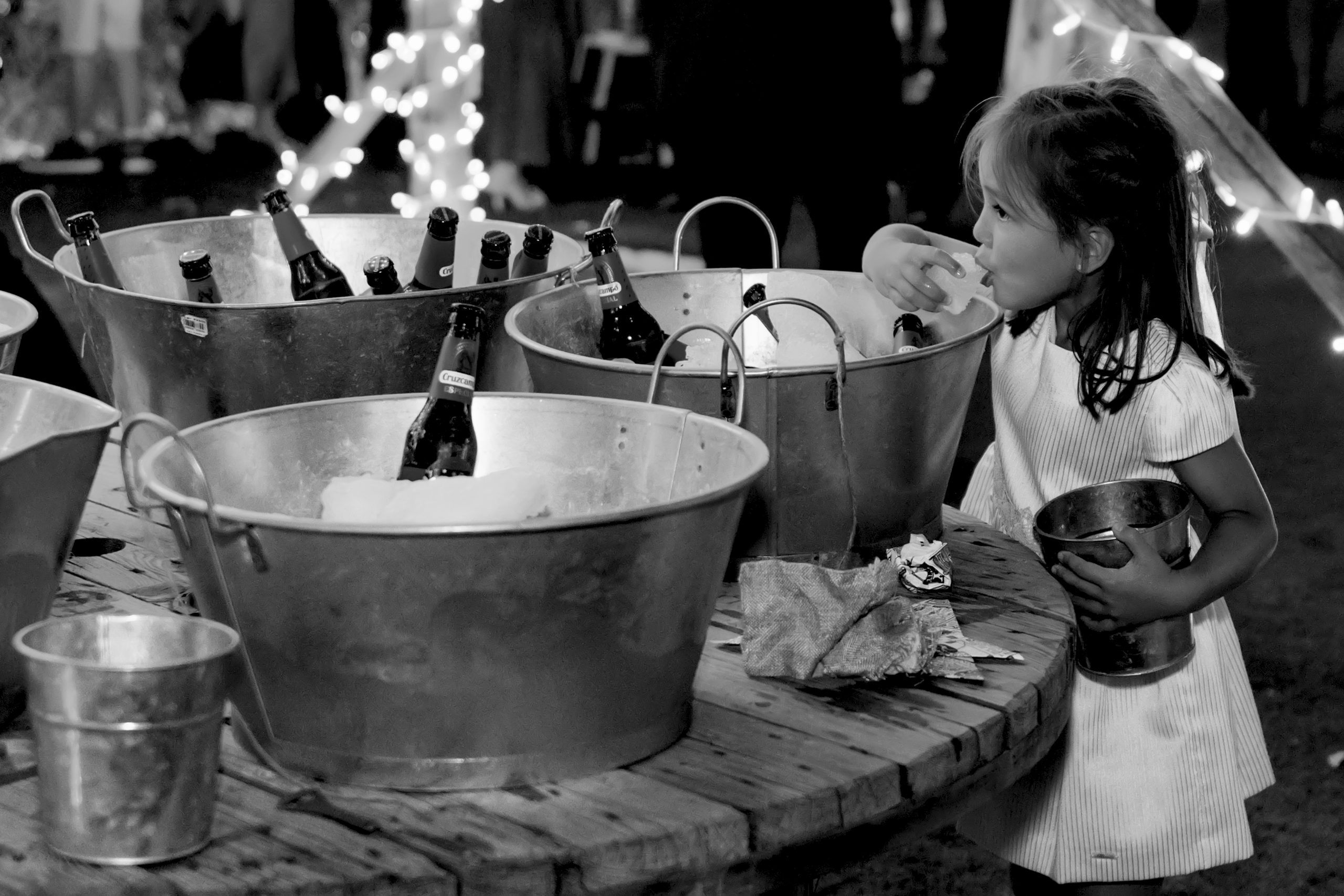 Foto en blanco y negro de una niña bebiendo de un cubito de hielo que ha cogido de los barreños donde se mantiene fría la bebida