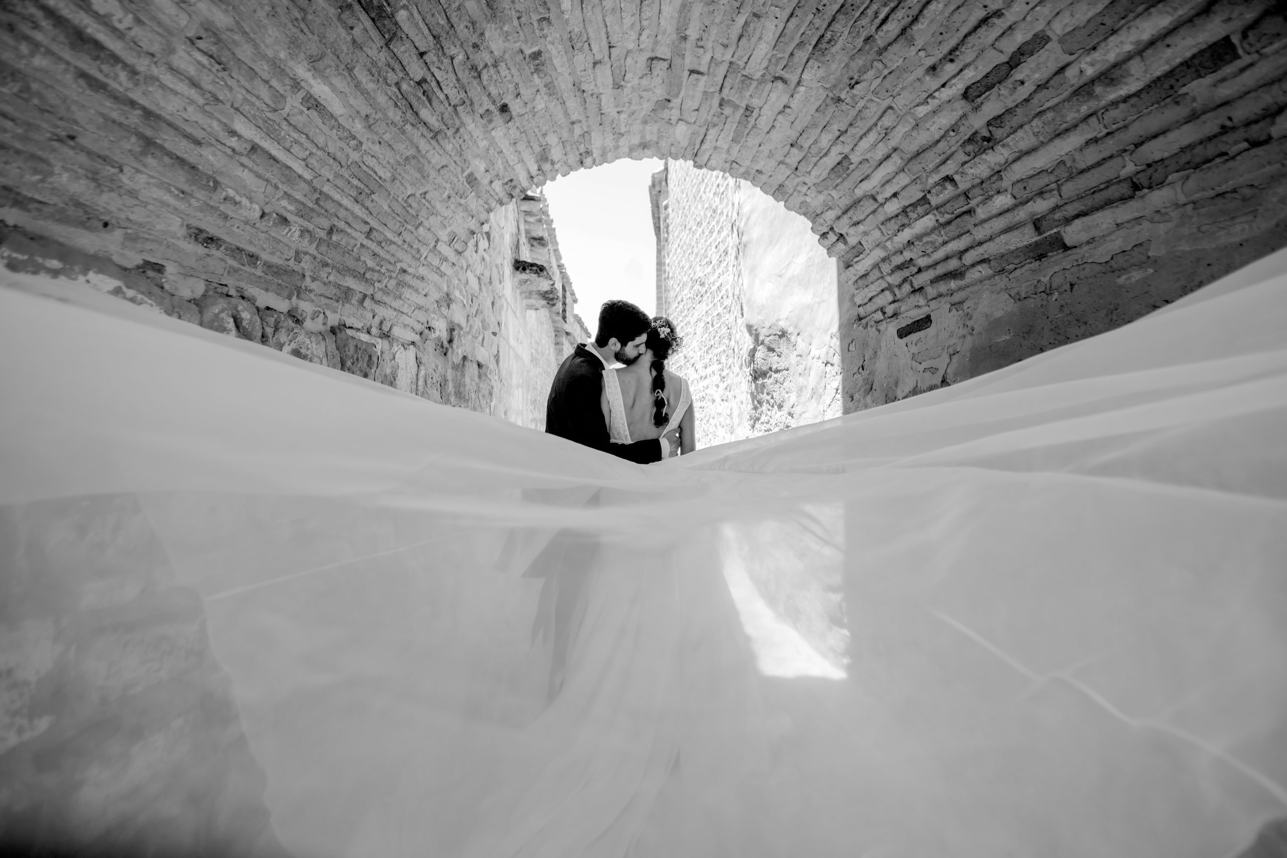 Foto en blanco y negro del novio besando a la novia en el cuello debajo de un arco de piedra enladrillado