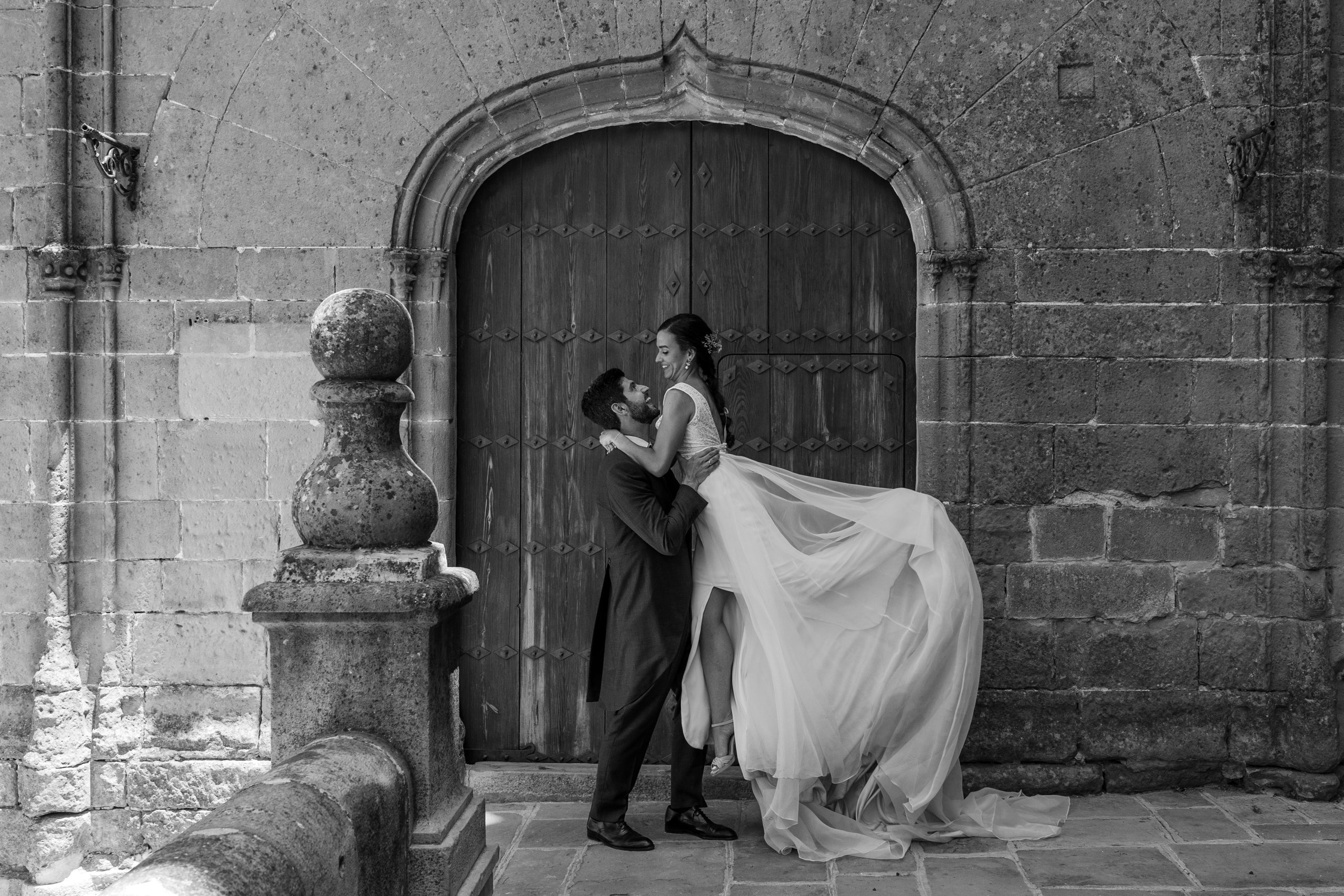 Foto en blanco y negro del novio elevando a la novia por encima del pecho y sonriendo con una puerta antigua detrás