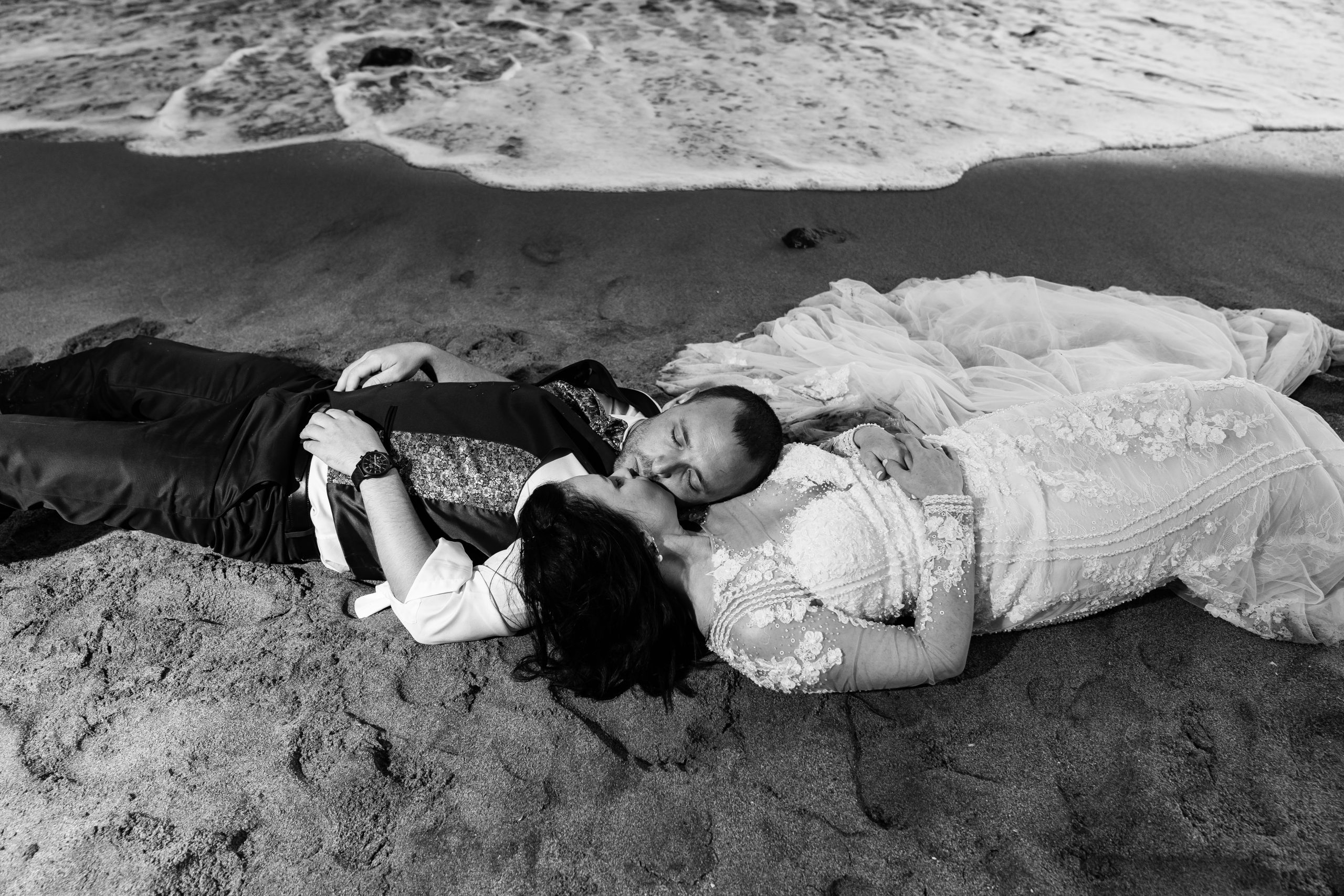 Foto en blanco y negro de los novios tumbados en la orilla del mar besándose