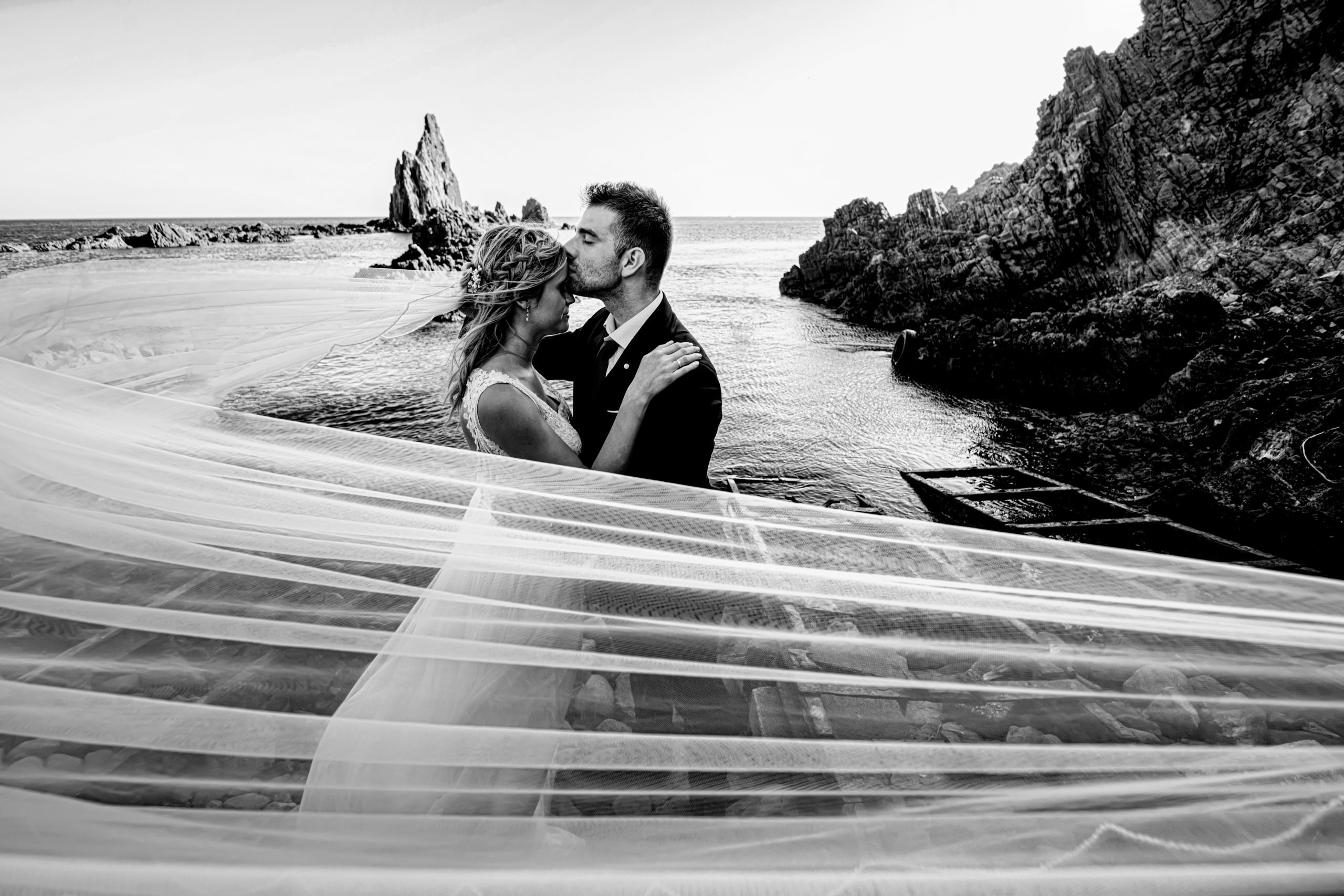 Foto en blanco y negro del novio besando la frente de la novia en la orilla del mar mientras el velo vuela
