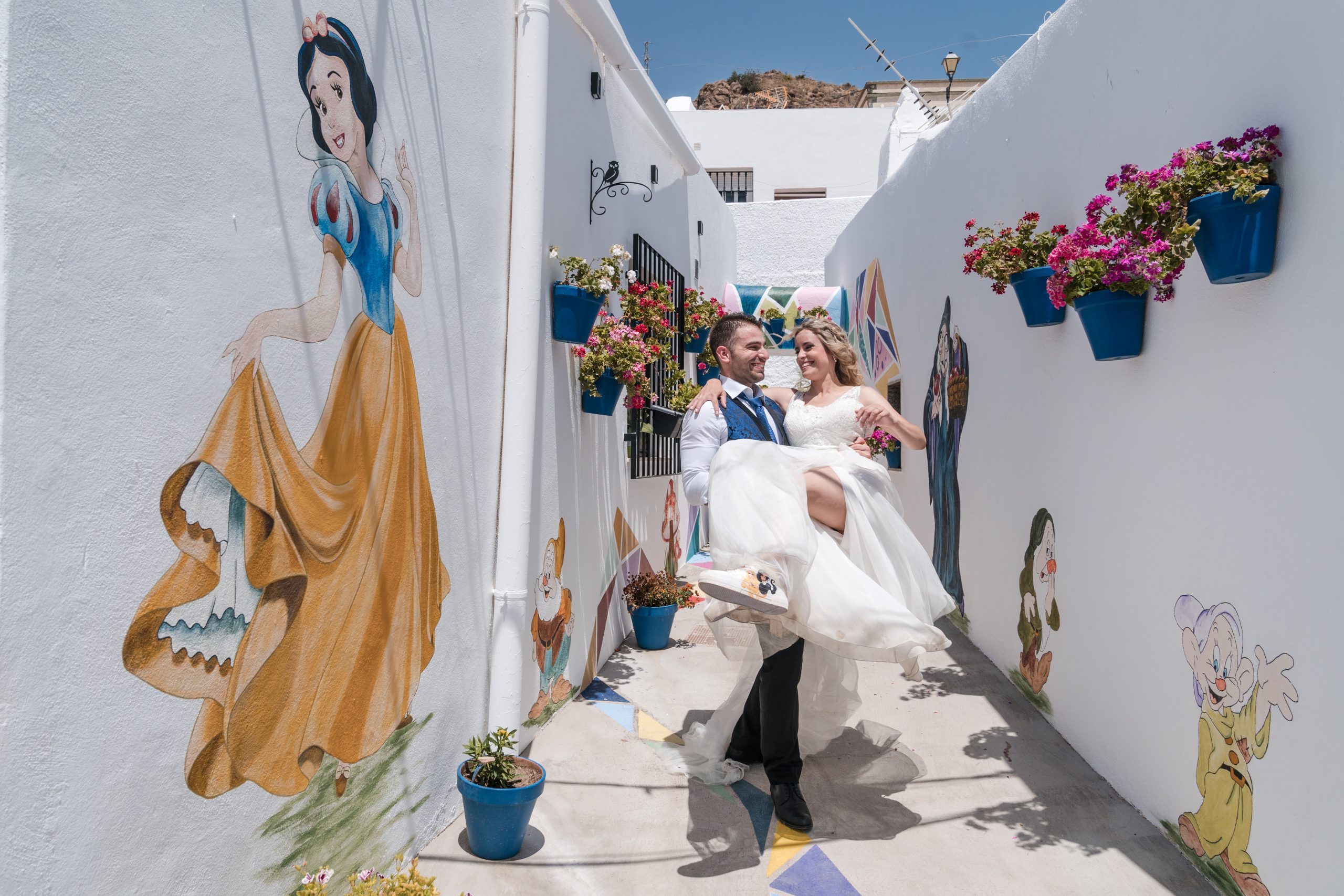 Novio llega en brazos a la novia por una calle blanca con muros pintados de personajes disney