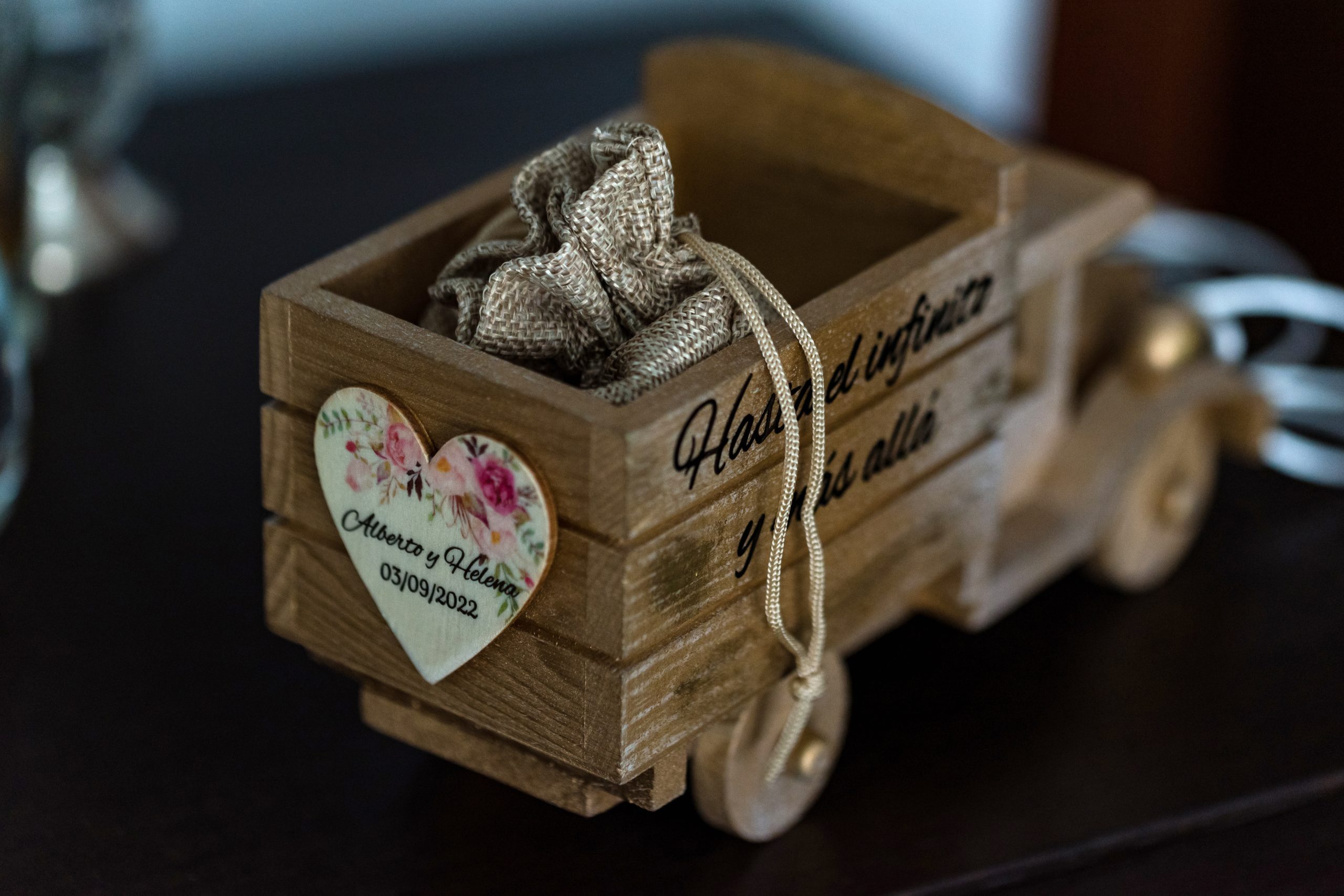 Un regalo de boda para los invitados de un camión de madera con una bolsita de tela en su interior, el camión tiene la fecha tres de septiembre de dos mil veintidós y los nombres Alberto y Helena atrás en una pegatina de forma de corazón