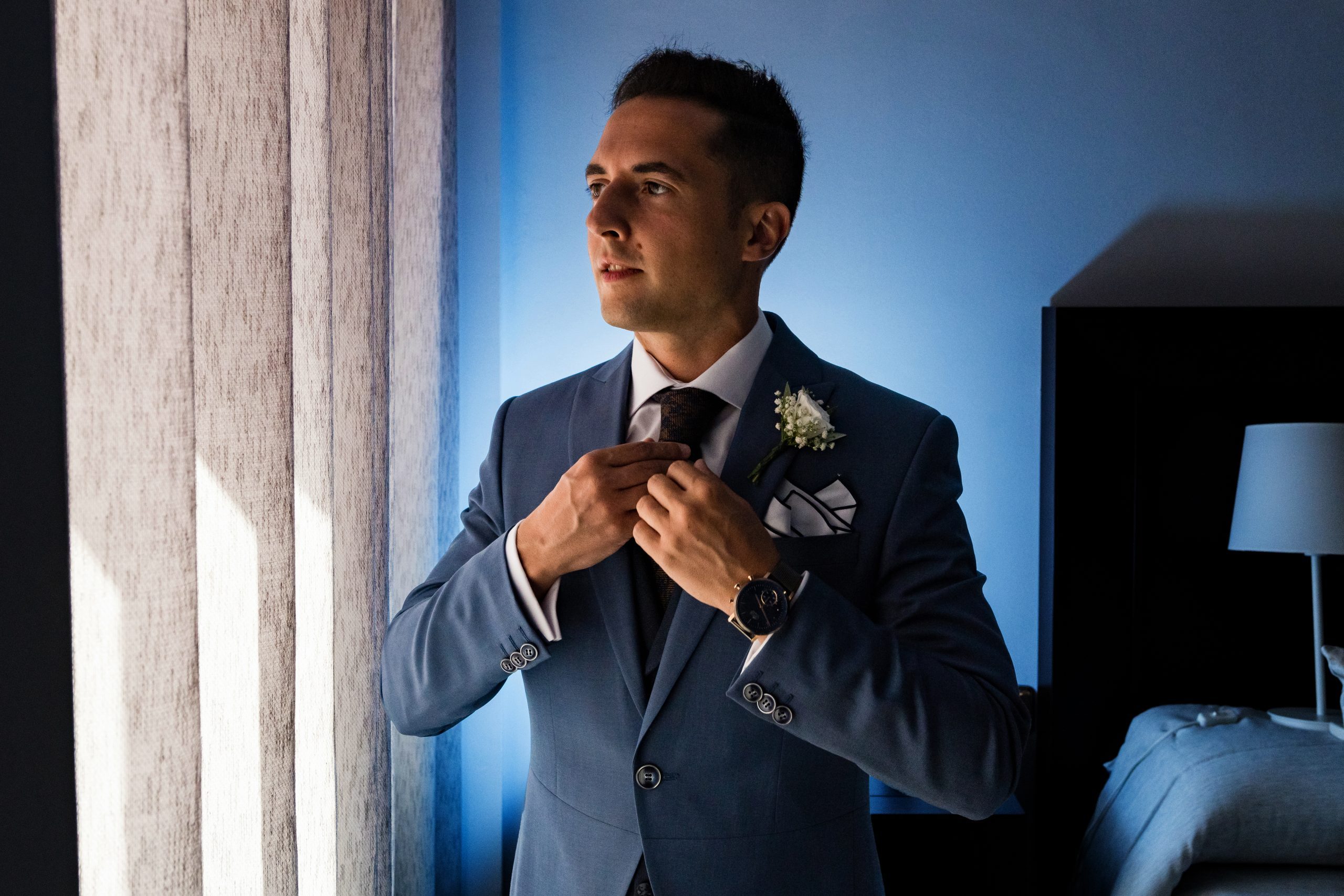 Novio mirando por la ventana colocándose la corbata antes de la boda