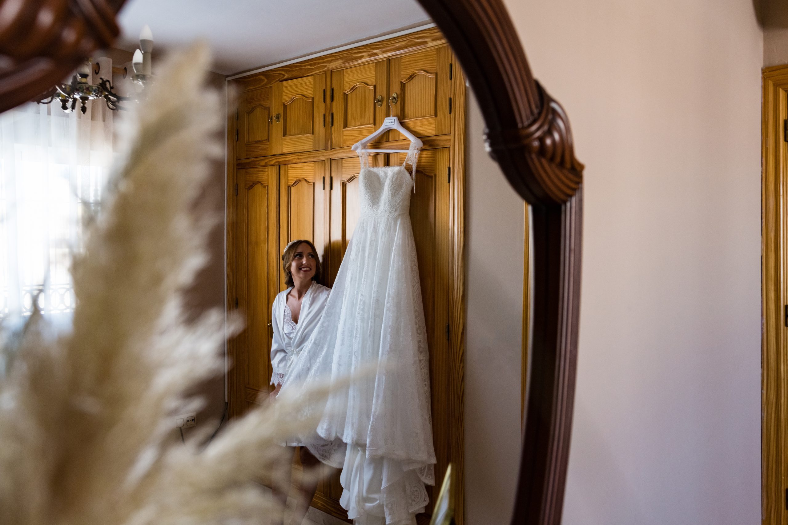 Foto de la novia alegre reflejada en el espejo mirando su vestido colgado del armario